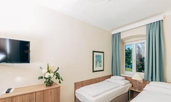 A bright room with two single beds and a television on the wall. Next to the window is a table with a flower decoration.