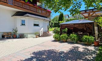 A cozy courtyard with a paved floor and an awning. In the background, there is a bright house with a balcony and blooming plants.