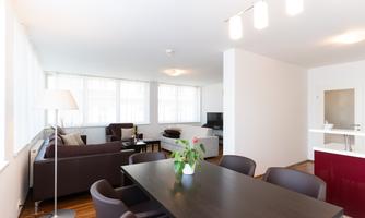 A bright and modern apartment with an open living and dining area. In the background, there is a sofa, a TV, and large windows that let in plenty of daylight.