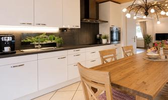 A modern kitchen with white cabinets and a large wooden table. The room has a warm atmosphere due to the wooden ceiling and the cozy decor.
