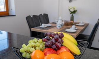 A fruit bowl with grapes, apples, oranges, and bananas is on the table. In the background, there is a dining area and a vase with flowers.