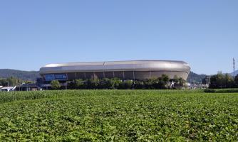 A modern stadium surrounded by green fields. In the background, gentle hills are visible.