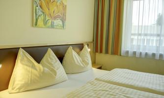A well-equipped room with two beds and cozy, decorative cushions. The walls are painted light and there is a colorful curtain and a piece of artwork.