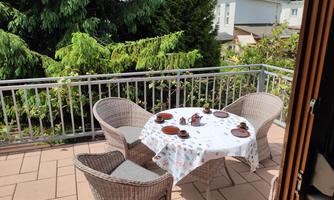 A cozy balcony with two rattan chairs and a set table. Surrounded by green plants, one observes a peaceful environment.