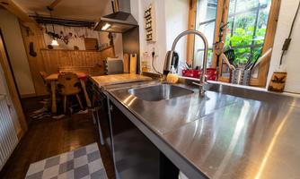 A modern kitchen with a stainless steel sink and wooden shelves. In the background, a dining area with a table and chairs is visible.