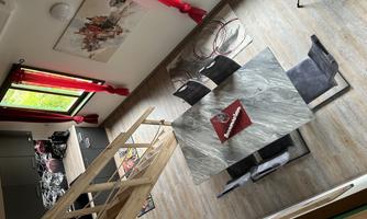 A modern room with a dining table and chairs, surrounded by large windows with red curtains. The kitchen is well-equipped and the floor is laid with light wood.