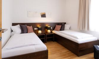 A cozy guest room with two single beds and a warm wooden floor. The walls are light and the decor is modern and simple.