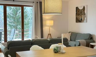 A cozy living room with a large sofa and a dining table. A picture of a wolf hangs on the wall and the windows offer a view of a snow-covered outdoor area.