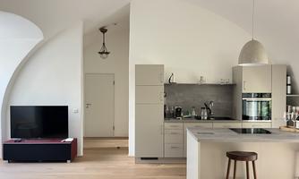 A modern kitchen with bright furnishings and an open layout. In the background, there is a television, and the room features large, curved walls.