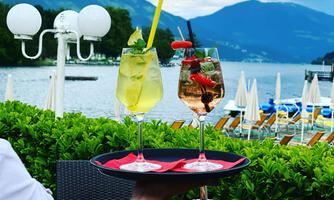 Two colorful cocktails on a tray in front of a picturesque lakeside landscape. In the background, loungers and sun umbrellas can be seen.