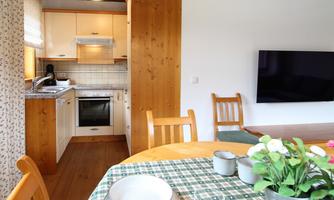 A cozy kitchen with a dining table and chairs. In the background, there is a TV and light wood details.