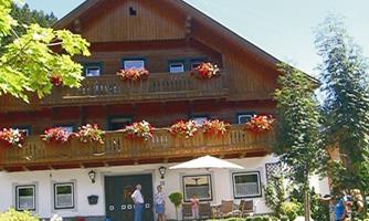 A traditional wooden house with blooming balcony boxes. People are standing in front of the entrance and enjoying the sunny surroundings.