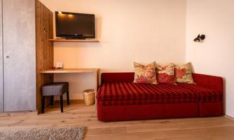 A cozy living area with a red sofa and decorative pillows. There is a table, a chair, and a television on the wall.