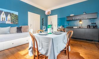 A cozy dining area with a round table and chairs. The walls are painted in a calming blue, and there is a modern kitchen unit in the background.