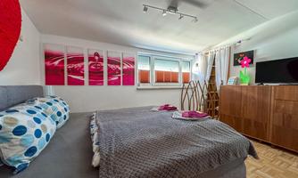 A modern bedroom with a large bed and colorful cushions. Pictures hang on the wall, and there is a television as well as a wooden wardrobe.