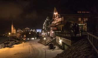A picturesque winter landscape at night with snow-covered streets and illuminated houses. In the background, a church is visible, emphasizing the tranquil atmosphere.