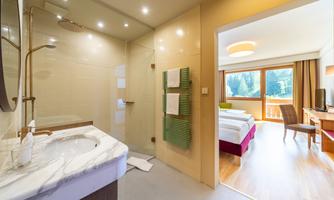 A modern bathroom with a shower and sink. In the background, a cozy bedroom with a view outside can be seen.