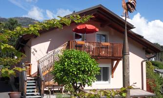 A cozy house with a balcony and a red parasol. In the foreground, there is a green tree and a weather vane.
