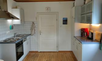 A modern kitchen with white cabinets and wooden flooring. It features a stove, oven, and a coffee machine.