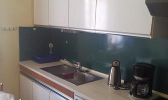 A simple kitchen with white cabinets and a green backsplash. The amenities include a sink, a kettle, and a coffee maker.