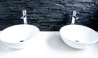 Two modern sinks made of white ceramic with chrome faucets. The backsplash is made of dark, textured stones.