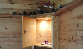 A cozy wooden room with an open cupboard. Inside, there is a bottle and a glass, surrounded by pinecones and decorative birds.