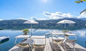 A picturesque lake with a wooden pier and sun umbrellas. In the background, there are hills and a clear blue sky.