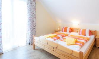 A bright bedroom with a large double bed and colorful cushions. The room has windows with curtains and a cozy wood look.