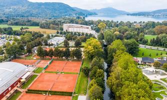 Aerial view shows tennis courts and lush trees in the surrounding area. In the background, mountains and a lake can be seen.