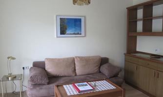 A cozy living room with a brown sofa, a wooden coffee table, and a bookshelf. A picture hangs on the wall, and the lighting is provided by a modern lamp.