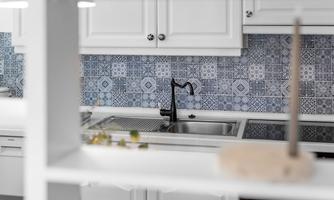 A modern kitchen with white cabinets and a blue patterned tile wall. The sink area is equipped with a stylish black faucet.