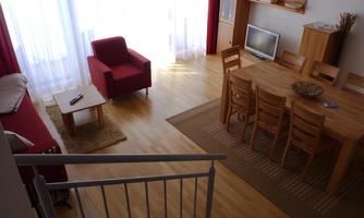A bright living room with wooden floors and large windows. There is a dining area with a wooden table and chairs, as well as a cozy seating area with a red armchair and a couch.