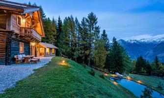 A cozy wooden house surrounded by trees and beautiful scenery. In the foreground, there is a small pond and the mountains are visible in the background.