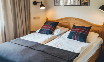 A cozy bedroom with a double bed and decorative cushions. Pictures hang on the walls, and the light is warm and inviting.