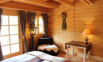 A cozy wooden room with a large window and curtains. A bed, a table, and a lamp complement the warm atmosphere.