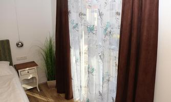 A cozy room with a bed and a nightstand. The windows are adorned with semi-transparent curtains featuring floral patterns.