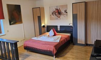 A cozy bedroom with a large bed and decorative pillows. The walls are decorated with modern artworks, and there is a television on a black cabinet.