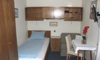 A simple room with a single bed, a table, and a chair. A television hangs on the wall and there are wooden furniture for storage.