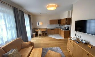 A cozy living room with a sofa, a wooden table, and a kitchenette. Bright curtains let in plenty of light and create a friendly atmosphere.