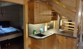 A cozy wooden kitchen with modern appliances and a small sink. In the background, a bedroom with a comfortable bed is visible.