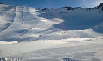 A snowy mountain landscape with freshly groomed ski slopes. The sky is clear and nature shines in its winter beauty.