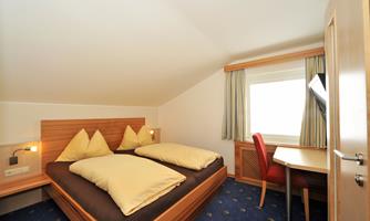 A cozy bedroom with a double bed and bright pillows. The room has a desk and a window with a view outside.