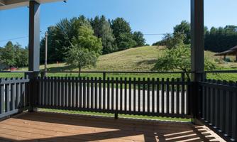 A view from a terrace with a wooden floor overlooking a green meadow and trees in the background. The sky is clear and sunny.