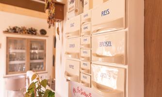 A cozy kitchen with a shelf of white compartments labeled with various foods. In the foreground, a green plant is growing, and a heart pendant is visible.
