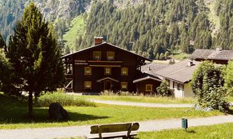 An idyllic alpine village with a traditional wooden house and green meadows. In the foreground, there is a park bench surrounded by trees.