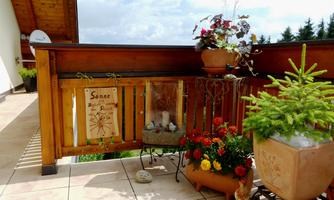 A beautiful balcony with blooming plants and a wooden railing. In the background, there is a wall decoration and a small water feature.