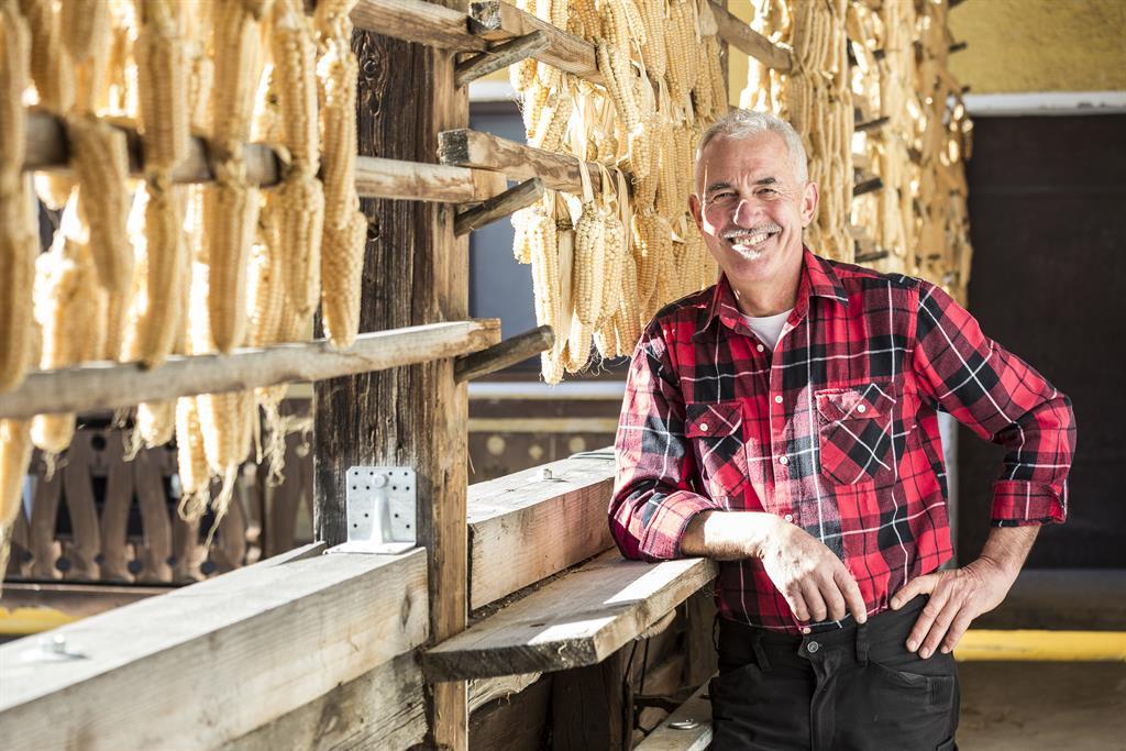 Bauer mit Maiskolben im Hintergrund lächelnd vor Holzwand