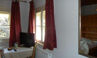 A simple room with a table, a chair, and a television. The windows are decorated with red curtains, and a bed is visible in the background.