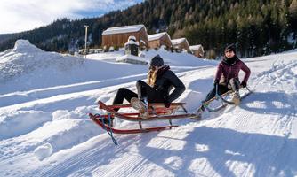Two people are riding on a sled in the snow. In the background, there are snow-covered hills and a wooden cabin.