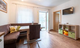 A cozy living area with a brown corner bench and a dining table. A television and a door lead to the balcony with a view.